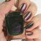 Hand model holding Cirque Colors Folie á Deux bottle, showcasing forest green magnetic nail polish with magenta-green-gold multichrome shimmer on nails